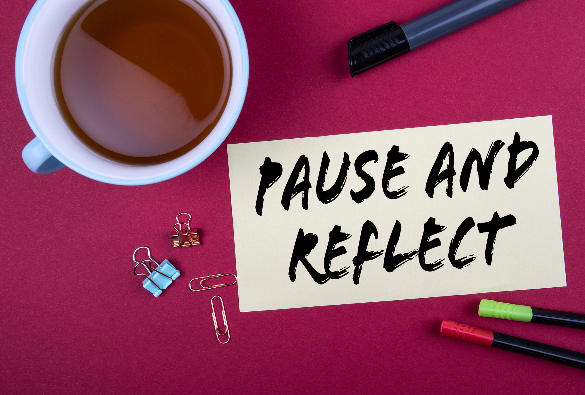 Sheet of paper with “Pause and Reflect” written on it beside a teacup and stationery on a red table, symbolizing reflection and intentional pause.